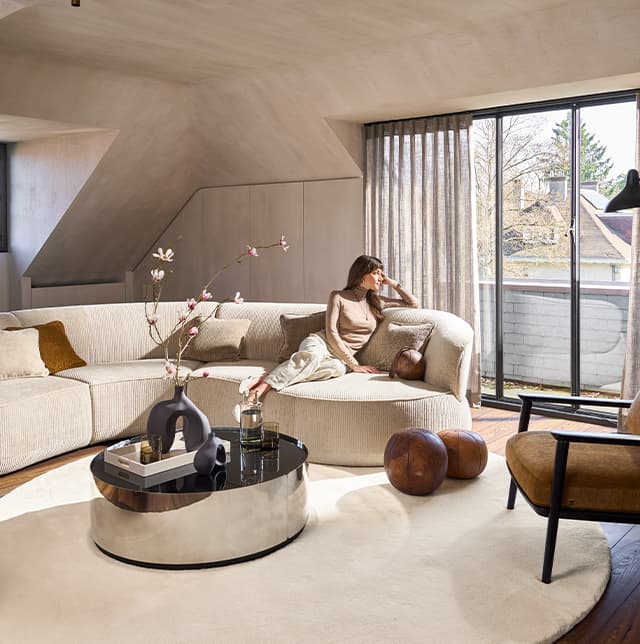 Eine Aufnahme von einem Wohnzimmer, gehalten in beige und braun. Eine Frau sitzt auf einem beigen Sofa und schaut in die Ferne.