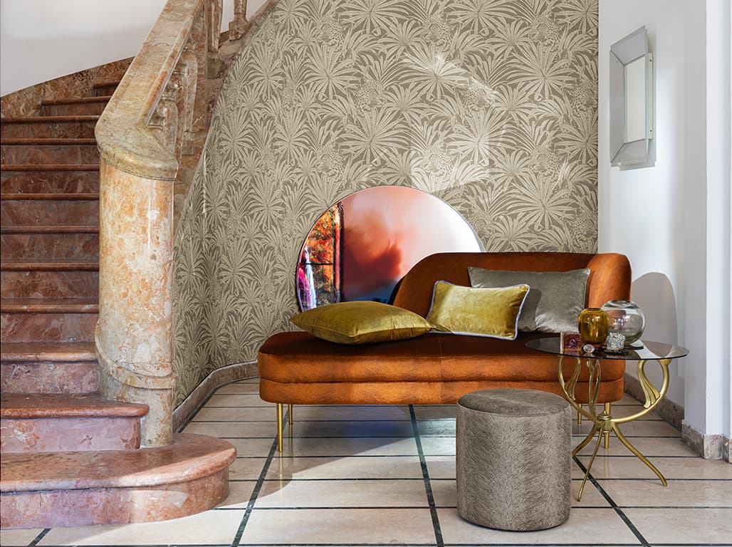 Das Bild zeigt eine braune Couch vor einer Treppe, dekoriert mit Kissen. Neben der Couch stehen ein grauer Pouf und ein Beistelltisch. Der Treppenaufgang ist mit einer Palmendrucktapete versehen, und die Farben harmonieren gut miteinander. 