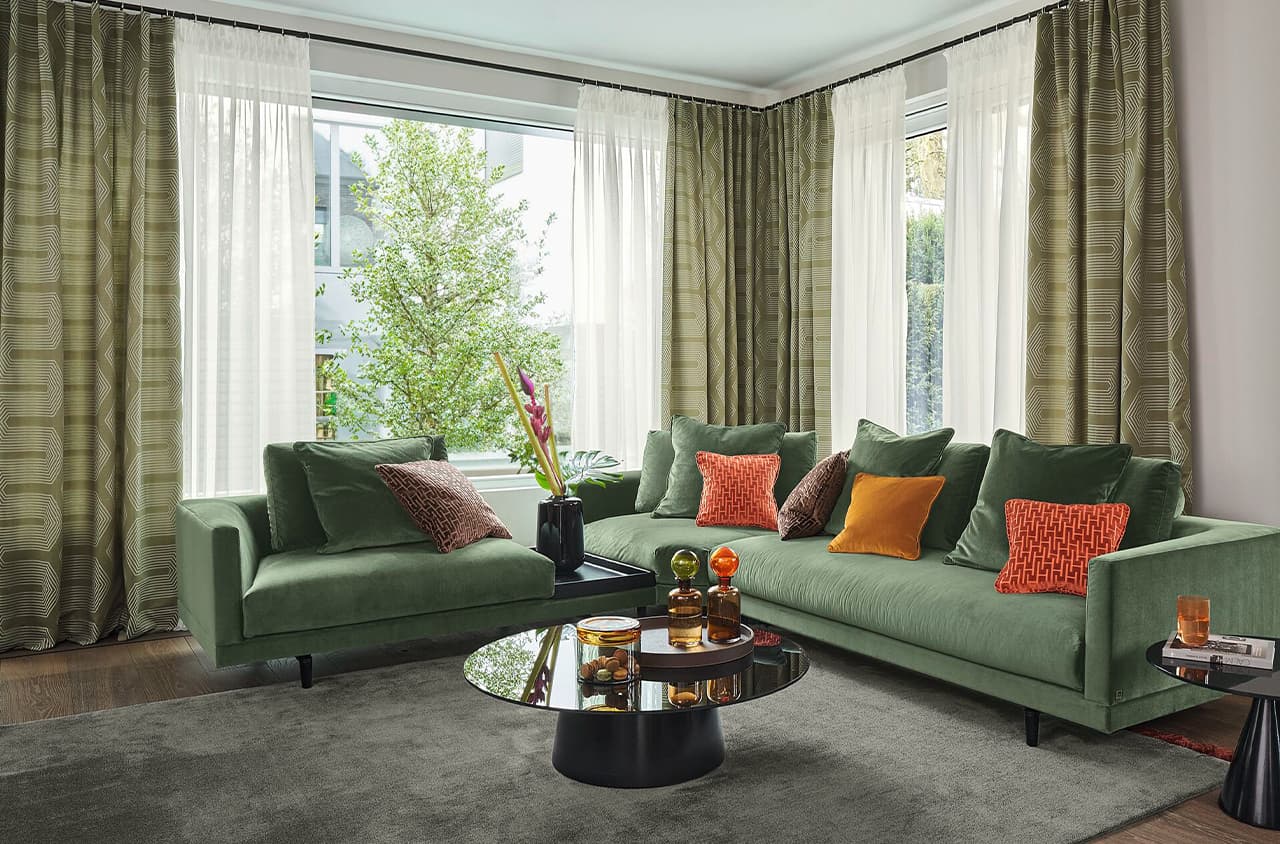 Ein grünes Sofa vor einer Fensterfront micht weißen und grünen Vorhängen. Im Raum sind orange und braune Akzente.