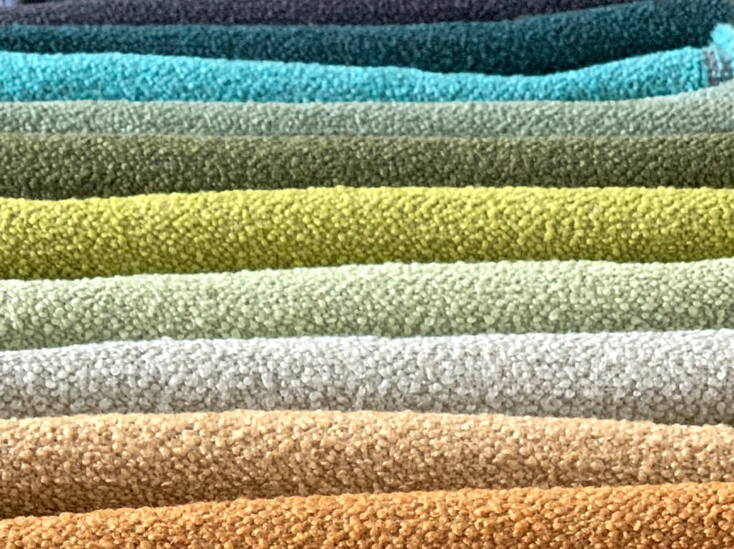 Das Bild zeigt eine Farbübersicht mit gestapelten Stoffen, die nach Farben sortiert sind. Die Farbpalette reicht von Braun über Grün bis Blau, wobei die Stoffe ordentlich angeordnet sind und eine harmonische Farbverlauf darstellen.