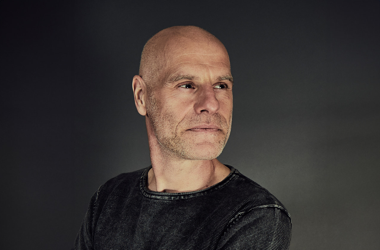Portraitfoto von einem Mann mit Glatze, er trägt einen grauen Pullover und schaut zur Seite.
