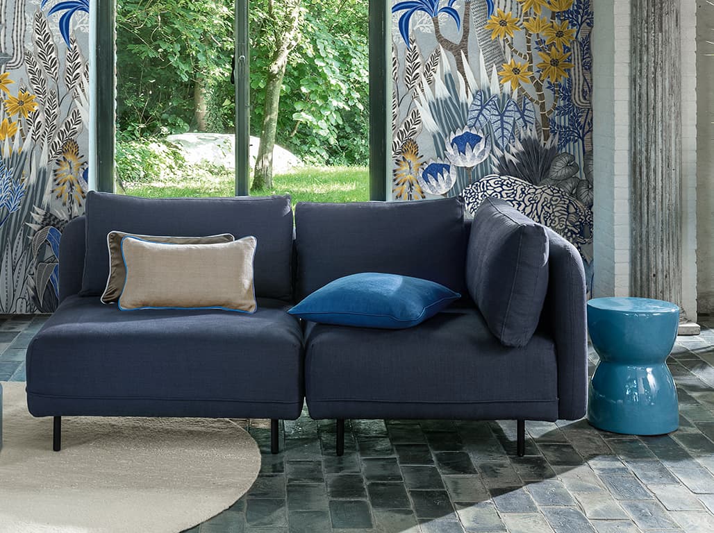 Blaues Sofa mit Kissen in grau und blau vor einer Tapete mit floralem Print.