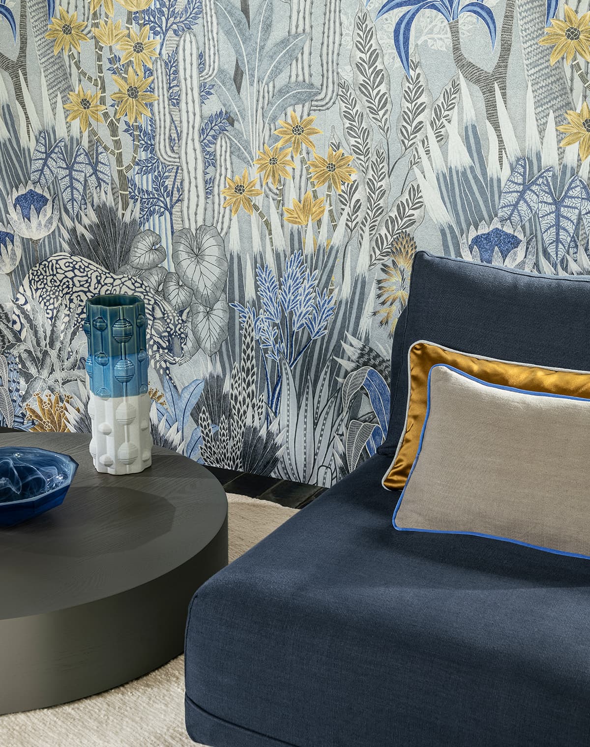Blaues Sofa mit einem weißen und einem goldenen Kissen vor einer Tapete mit floralem Print.
