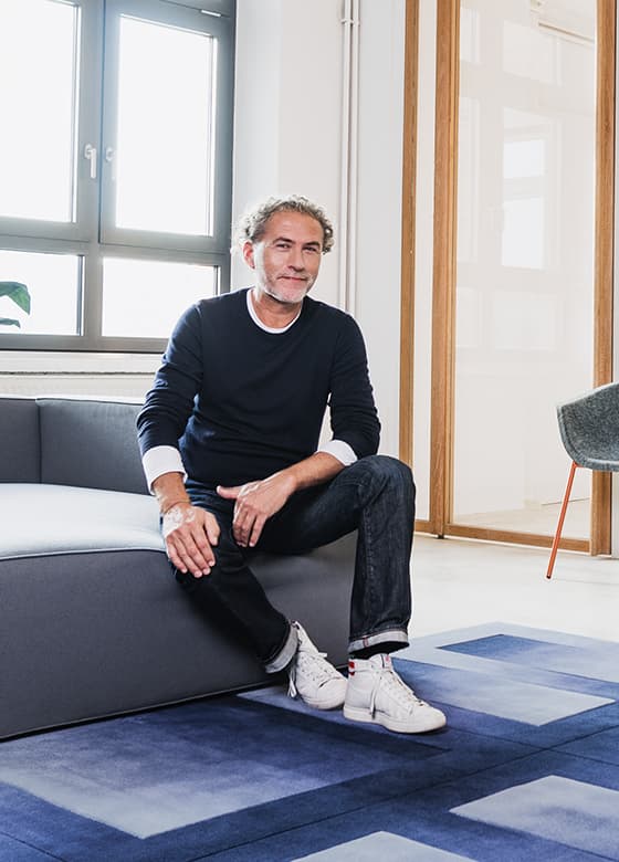Produktdesigner Werner Aisslinger sitz auf einer grauen Couch welche auf einem blauen Teppich steht
