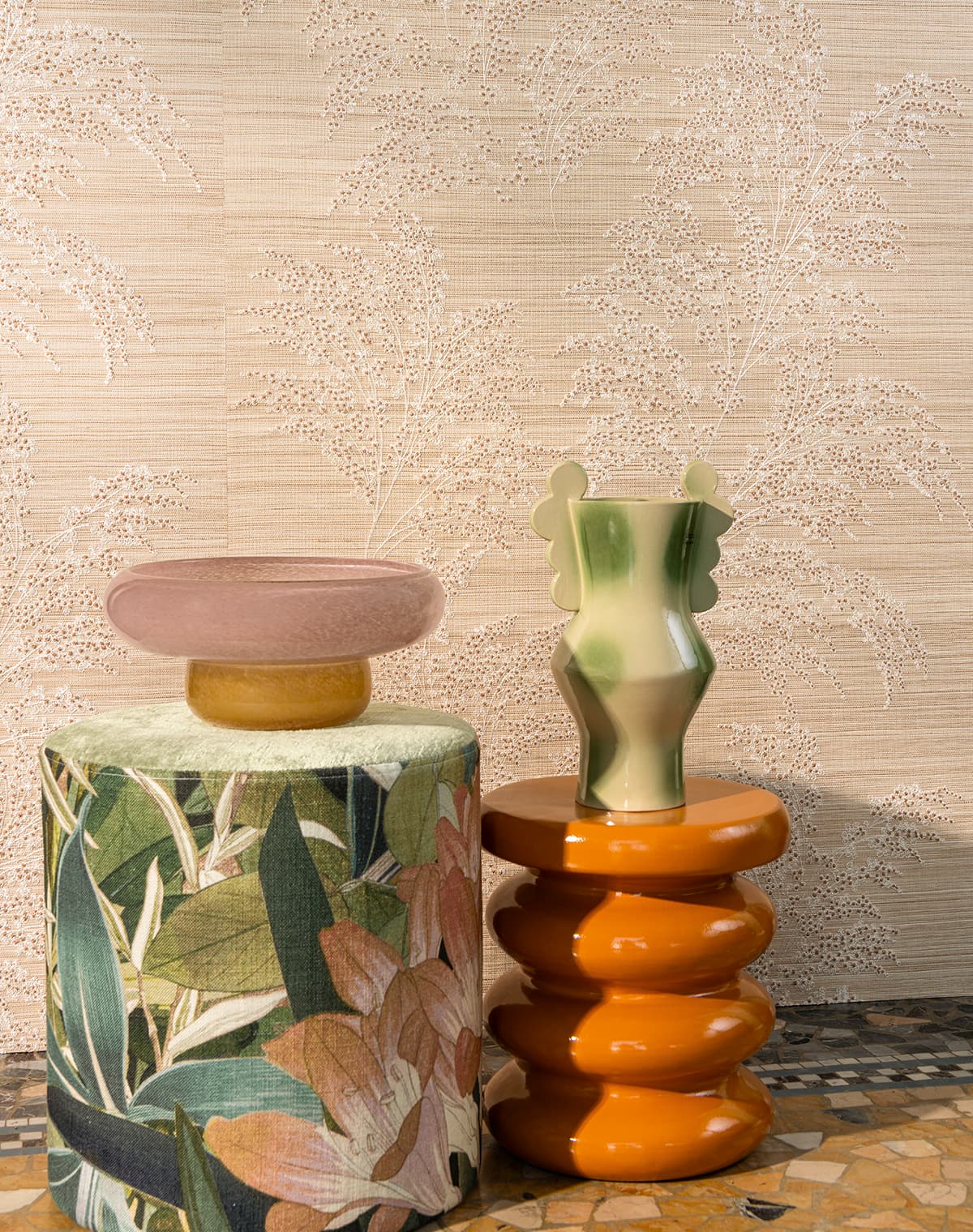Tapete mit floralem Muster hinter einem Pouf mit floralem Muster, einem orangenen Beistelltisch und einer grünen Vase.