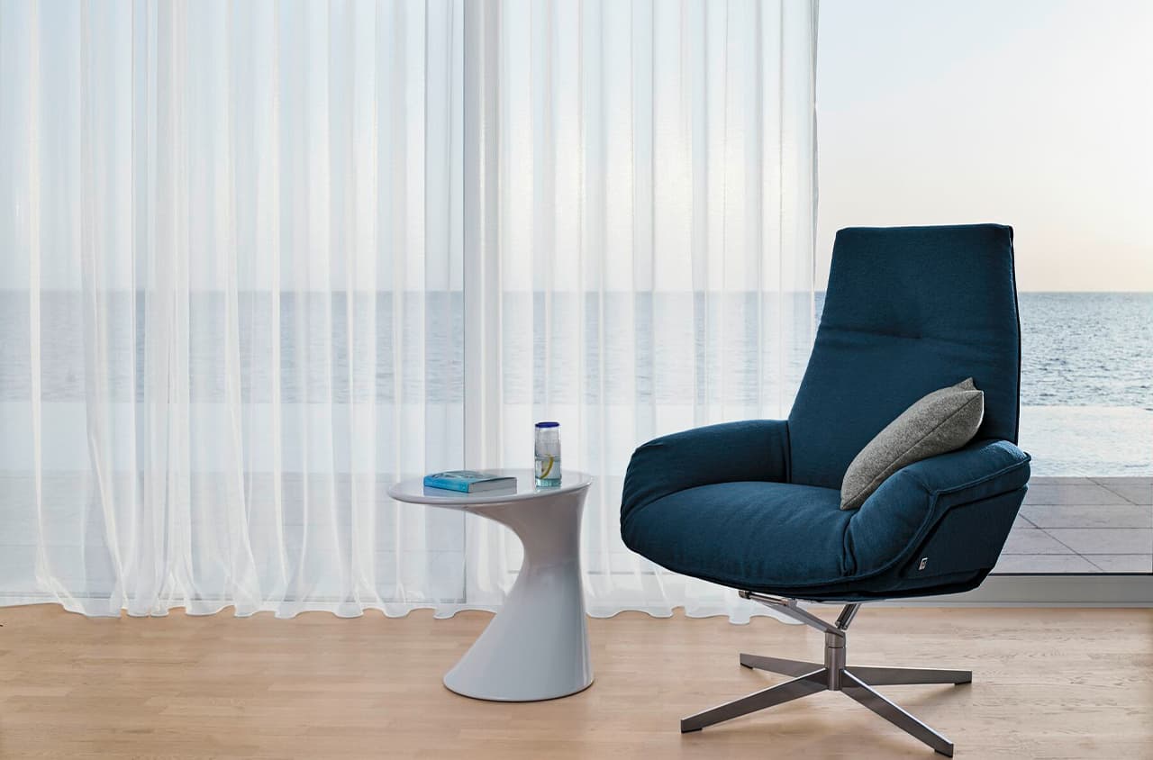 Ein blauer Sessel und ein weißer Beistelltisch stehen vor einem Fenster mit einem durchsichtigen Vorhang.